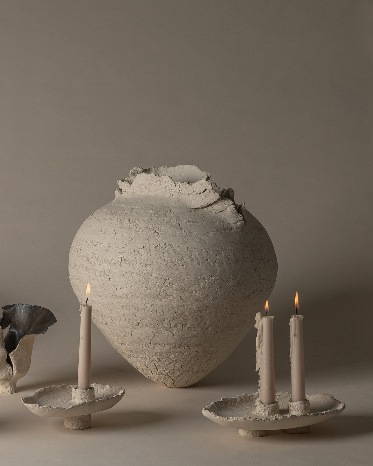 Powder single and double candlesticks holding lit candles, with a ceramic vessel behind them. Each candlestick is sold individually.